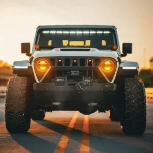 Gray Jeep Wrangler JL with jeep gladiator headlights glowing amber warm light driving on road at sunset golden hour.