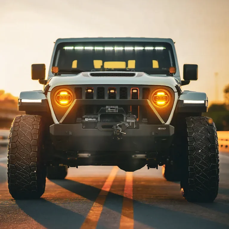 Gray Jeep Wrangler JL with jeep gladiator headlights glowing amber warm light driving on road at sunset golden hour.