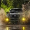 Black Toyota Tacoma driving through muddy water with two yellow fog lights visible through the spray