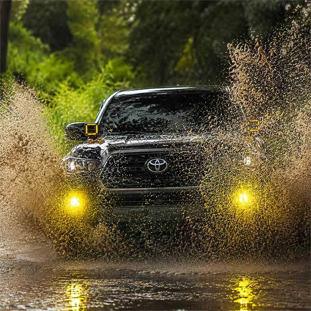 Black Toyota Tacoma driving through muddy water with two yellow fog lights visible through the spray