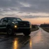Dark green Toyota Tacoma parked beside a wet rural road at dusk with both yellow fog lights reflecting on the pavement