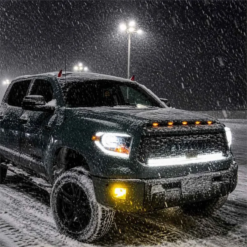 Dark Toyota Tundra parked in heavy snow with bright white headlights amber marker lights and yellow bumper fog lights