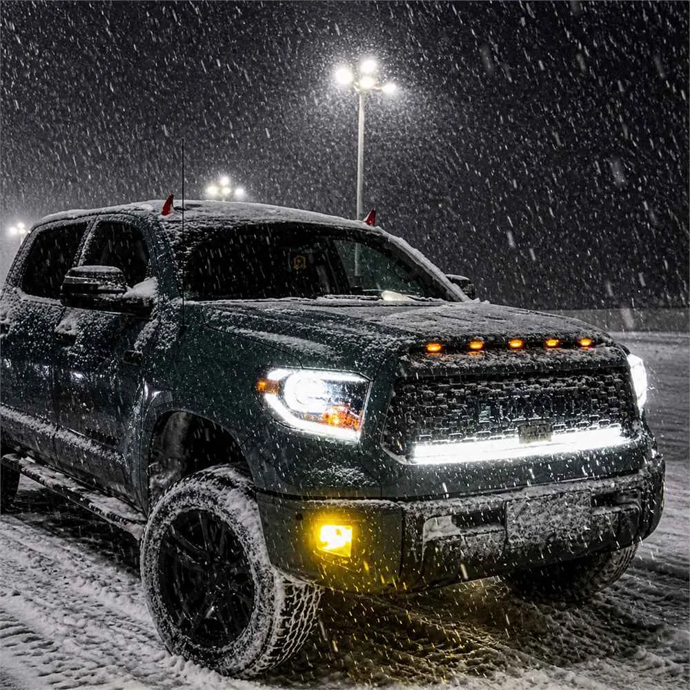 Dark Toyota Tundra parked in heavy snow with bright white headlights amber marker lights and yellow bumper fog lights