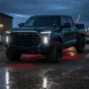 Three-quarter view of a dark Toyota Tundra on wet pavement with white vent DRLs and headlights illuminated