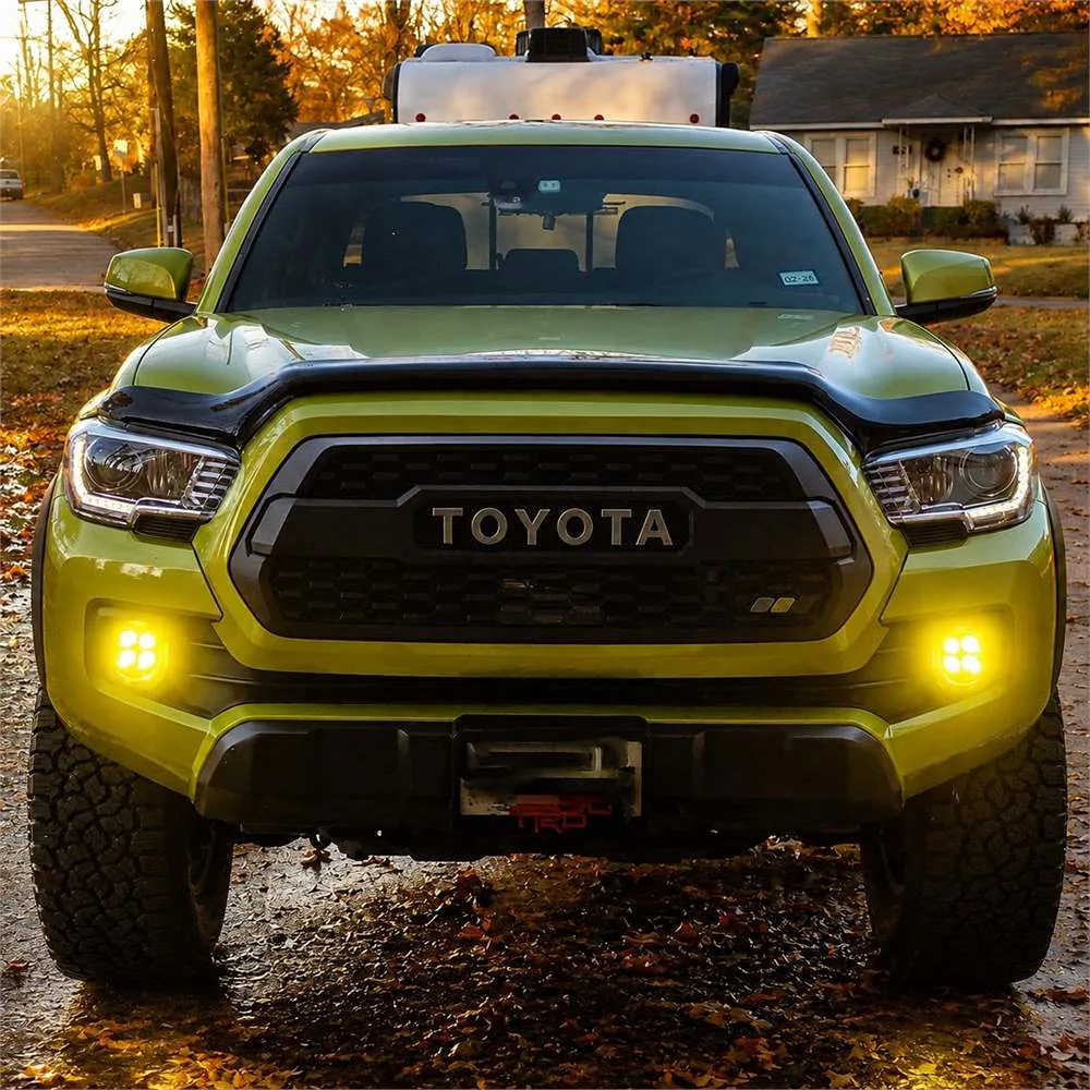 Front view of a green TRD-style Toyota Tacoma with both yellow bumper fog lights illuminated in autumn surroundings